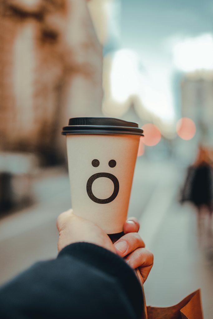 A disposable coffee cup with a playful face held on a city street, featuring a blurred background.